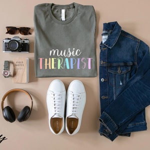 May include: A flat lay of a green t-shirt with the text "music therapist" in rainbow colors, a pair of white sneakers, a denim jacket, a camera, a watch, a notebook, and a pair of headphones. The word "Army" is written in black cursive in the bottom left corner of the image.