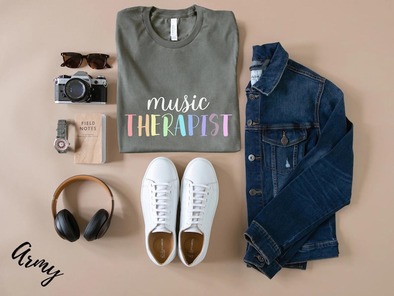 May include: A flat lay of a green t-shirt with the text "music therapist" in rainbow colors, a pair of white sneakers, a denim jacket, a camera, a watch, a notebook, and a pair of headphones. The word "Army" is written in black cursive in the bottom left corner of the image.
