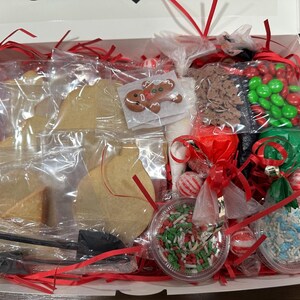 May include: A white box filled with a cookie decorating kit. The kit includes various cookie shapes, icing, sprinkles, candies, and a gingerbread man cookie. Red ribbon and cellophane wrap the contents.