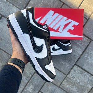 May include: Black and white Nike low-top sneakers with a black swoosh and black rhinestones. The sneakers are on a gray brick surface with a red Nike box in the background.