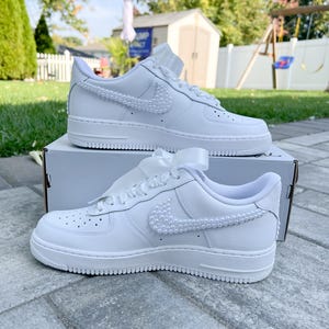 K&ouml;nnte beinhalten: Wei&szlig;e Ledersneaker mit einem mit Perlen besetzten Nike-Swoosh. Die Sneaker sind mit einem wei&szlig;en Satinband gebunden und stehen auf einem wei&szlig;en Karton.