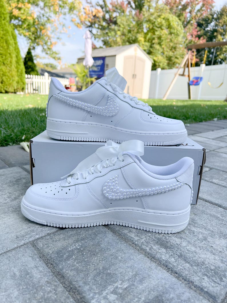 K&ouml;nnte beinhalten: Wei&szlig;e Ledersneaker mit einem mit Perlen besetzten Nike-Swoosh. Die Sneaker sind mit einem wei&szlig;en Satinband gebunden und stehen auf einem wei&szlig;en Karton.
