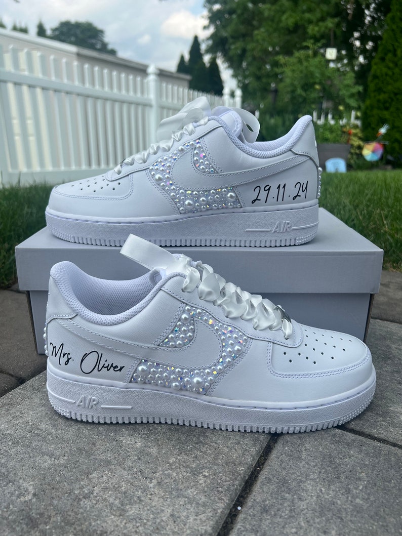 Puede incluir: Zapatillas blancas de cuero con un dise&ntilde;o de perlas y diamantes de imitaci&oacute;n en el swoosh. Las zapatillas tienen una cinta blanca atada alrededor y el texto "Mrs. Oliver" y "29.11.24" escrito en ellas.