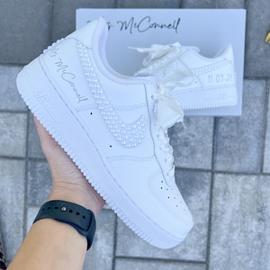 K&ouml;nnte beinhalten: Wei&szlig;e Lederschuhe mit einer Perlenverzierung am Swoosh und an der Seite. Die Schuhe haben ein wei&szlig;es Satinband, das um die Schn&uuml;rsenkel gebunden ist, und der Text "Mrs. McConnell" ist an der Seite geschrieben. Das Datum "11.09.20" ist auf dem anderen Schuh geschrieben.