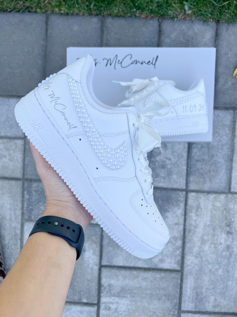 K&ouml;nnte beinhalten: Wei&szlig;e Lederschuhe mit einer Perlenverzierung am Swoosh und an der Seite. Die Schuhe haben ein wei&szlig;es Satinband, das um die Schn&uuml;rsenkel gebunden ist, und der Text "Mrs. McConnell" ist an der Seite geschrieben. Das Datum "11.09.20" ist auf dem anderen Schuh geschrieben.
