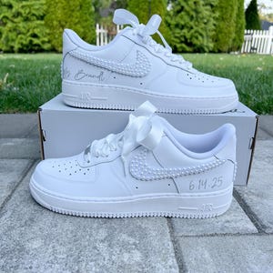 K&ouml;nnte beinhalten: Wei&szlig;e Lederschuhe mit einem wei&szlig;en Satinband, das um die Schn&uuml;rsenkel gebunden ist. Die Schuhe haben einen mit Perlen besetzten Nike-Swoosh und den Text "Mrs. Brandt" und "6.14.25" darauf geschrieben.
