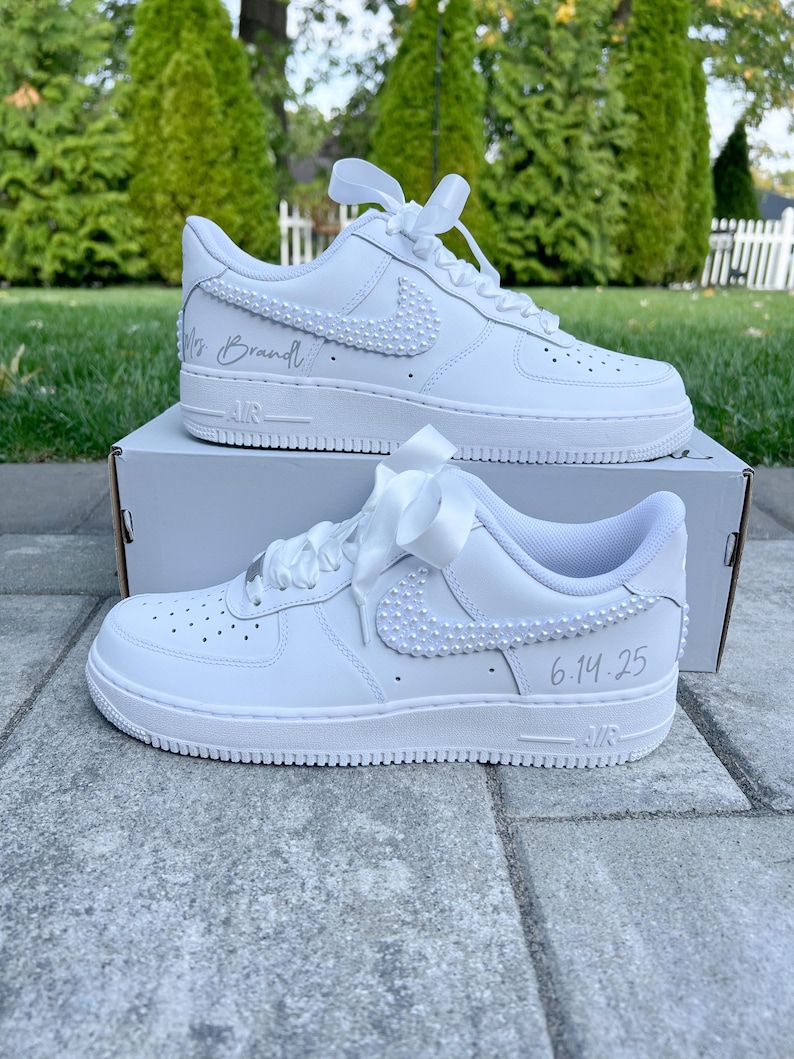K&ouml;nnte beinhalten: Wei&szlig;e Lederschuhe mit einem wei&szlig;en Satinband, das um die Schn&uuml;rsenkel gebunden ist. Die Schuhe haben einen mit Perlen besetzten Nike-Swoosh und den Text "Mrs. Brandt" und "6.14.25" darauf geschrieben.