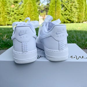 K&ouml;nnte beinhalten: Ein Paar wei&szlig;e Nike Air Force 1 Sneaker mit wei&szlig;en Satinb&auml;ndern, die um die Kn&ouml;chel gebunden sind. Die Sneaker haben eine Reihe wei&szlig;er Perlen an der Seite und die Worte "Nike Air" sind auf der R&uuml;ckseite jedes Schuhs aufgedruckt.