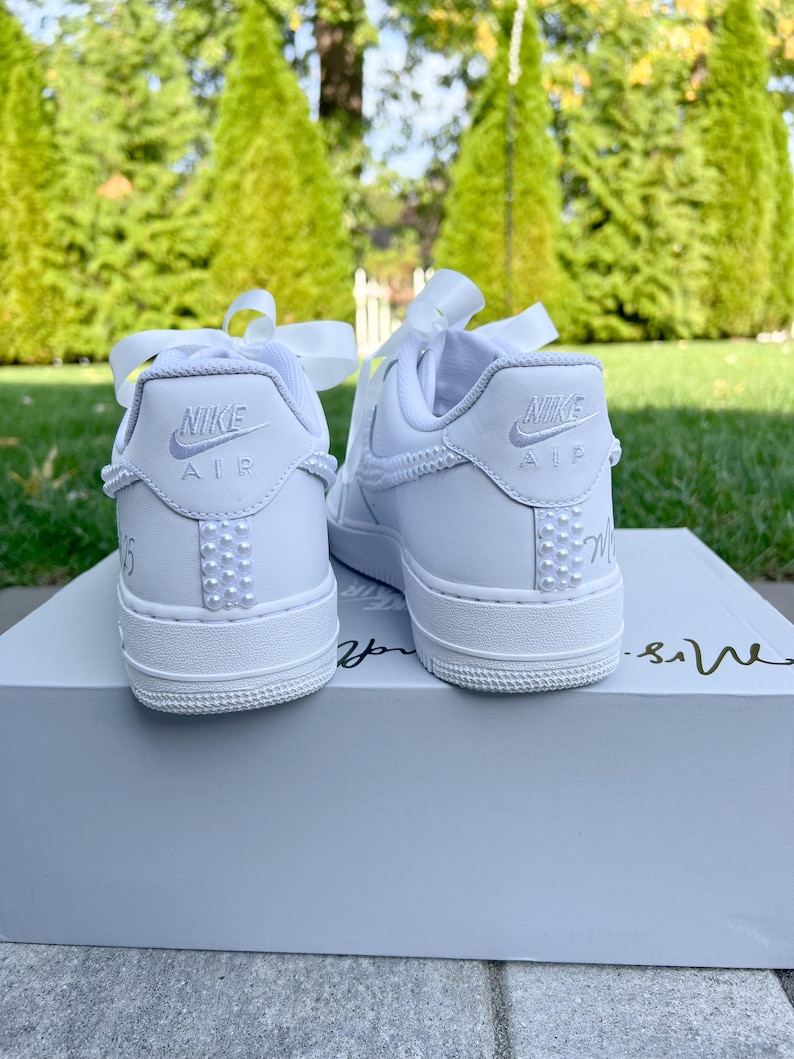 K&ouml;nnte beinhalten: Ein Paar wei&szlig;e Nike Air Force 1 Sneaker mit wei&szlig;en Satinb&auml;ndern, die um die Kn&ouml;chel gebunden sind. Die Sneaker haben eine Reihe wei&szlig;er Perlen an der Seite und die Worte "Nike Air" sind auf der R&uuml;ckseite jedes Schuhs aufgedruckt.