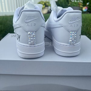 Puede incluir: Un par de zapatillas Nike Air Force 1 de cuero blanco con adornos de perlas en la parte posterior. Las zapatillas est&aacute;n sobre una caja blanca con el logotipo de Nike Air Force 1.