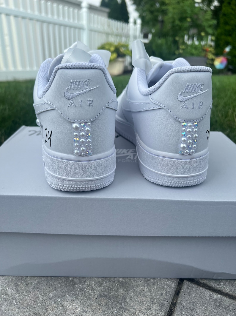 Puede incluir: Un par de zapatillas Nike Air Force 1 de cuero blanco con adornos de perlas en la parte posterior. Las zapatillas est&aacute;n sobre una caja blanca con el logotipo de Nike Air Force 1.