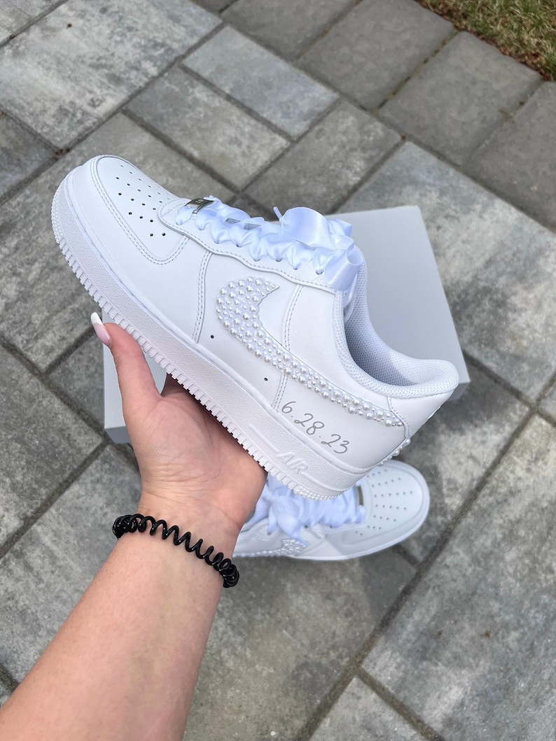 K&ouml;nnte beinhalten: Wei&szlig;e Lederschuhe mit einem wei&szlig;en Satinband, das um die Schn&uuml;rsenkel gebunden ist. Die Schuhe haben ein Perlendesign auf dem Swoosh und den Text "6.28.23" an der Seite.