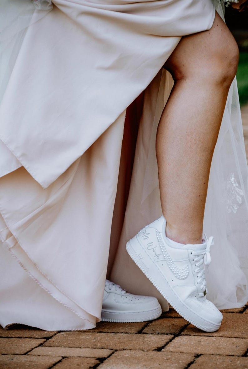 K&ouml;nnte beinhalten: Eine Braut in einem wei&szlig;en Hochzeitskleid und wei&szlig;en Turnschuhen mit dem Text "Mrs. Wonderful" auf einem Schuh.