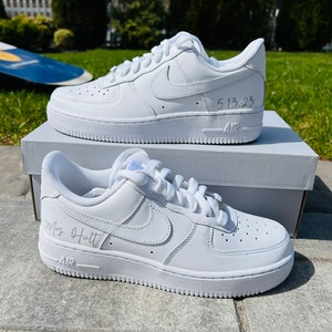 K&ouml;nnte beinhalten: Ein Paar wei&szlig;e Nike Air Force 1 Sneaker aus Leder mit einer personalisierten Inschrift auf jedem Schuh. Der linke Schuh sagt "5.13.23" und der rechte Schuh sagt "Mrs. Holt".