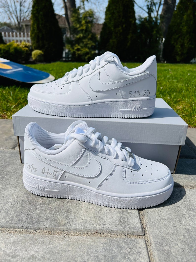K&ouml;nnte beinhalten: Ein Paar wei&szlig;e Nike Air Force 1 Sneaker aus Leder mit einer personalisierten Inschrift auf jedem Schuh. Der linke Schuh sagt "5.13.23" und der rechte Schuh sagt "Mrs. Holt".