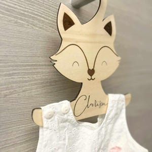 Può includere: Un appendiabiti di legno a forma di volpe con un vestito bianco appeso. L'appendiabiti ha il nome "Clarissa" inciso su di esso.