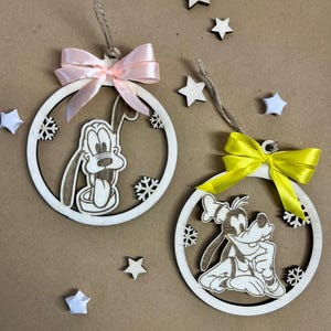 May include: Two wooden Christmas ornaments featuring Pluto, each with a different pose, are displayed. One ornament has a pink bow, the other a yellow bow. The ornaments are surrounded by small wooden stars and snowflakes.