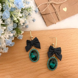 May include: A pair of drop earrings featuring a gold-tone hook, a black fabric bow, and an oval-shaped dark green faceted gemstone surrounded by a border of small, bright blue crystals. The earrings are displayed on a smooth, light-colored wooden surface.