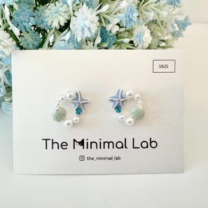 May include: A pair of silver earrings with a blue starfish, a green seashell, and white pearls. The earrings are on a white card with the text "The Minimal Lab" and "@the_minimal_lab".