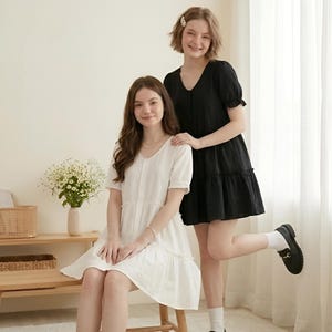 May include: A white short-sleeved tiered dress with a V-neckline and a black short-sleeved tiered dress with a V-neckline. Both dresses feature a button-down front, gathered waist seams, and ruffled hem details, presented as a matching set.