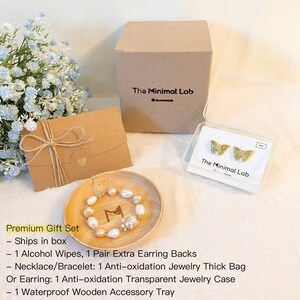 Puede incluir: Un conjunto de regalo premium para joyer&iacute;a, que incluye una bandeja de madera, un estuche de joyer&iacute;a transparente, un sobre marr&oacute;n con un coraz&oacute;n y una caja con el texto "The Minimal Lab". El conjunto incluye un collar o pulsera de perlas, un par de aretes de mariposa y respaldos de aretes adicionales.