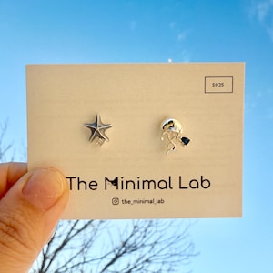 May include: A pair of silver stud earrings on a display card labeled The Minimal Lab. One earring is a starfish shape with a small clear stone, and the other is a jellyfish shape with a dark blue stone. The card includes the text S925.