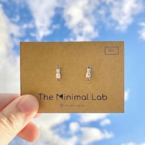 May include: Silver cat stud earrings presented on a brown card, with the text "The Minimal Lab" and the Instagram handle "@the_minimal_lab". The background is a blue sky with white clouds.