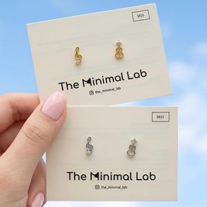 May include: Two pairs of stud earrings on display cards labeled The Minimal Lab. One pair is gold-toned with a treble clef and a guitar shape, each featuring a small clear stone. The second pair is silver-toned with identical musical motifs.