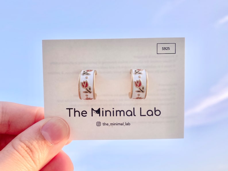 May include: A pair of white hoop earrings featuring a small red tulip floral pattern and gold-toned edges. The earrings are mounted on a display card labeled with the text The Minimal Lab and S925, with an Instagram handle icon and text.