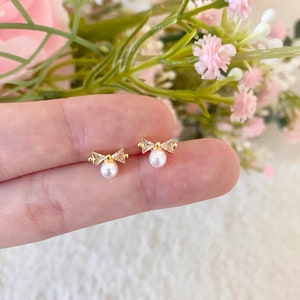 Może przedstawiać: Para złotych kolczyków w kształcie kokardki z małymi białymi perłami zwisającymi z dołu.