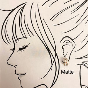 May include: A small hoop earring with a white base, gold-toned edges, and a delicate floral pattern featuring red and green details. The earring is displayed on a black and white line drawing of a profile, with the word Matte printed below.