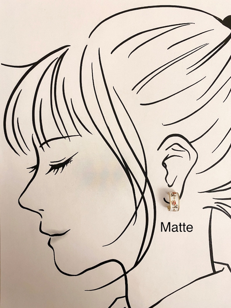 May include: A small hoop earring with a white base, gold-toned edges, and a delicate floral pattern featuring red and green details. The earring is displayed on a black and white line drawing of a profile, with the word Matte printed below.