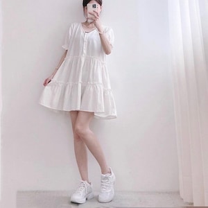 May include: A white, tiered, short-sleeve dress with a button-down front. The dress is paired with white sneakers. The model is standing in front of a white wall and a white curtain.
