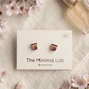 May include: A pair of stud earrings shaped like bubble tea cups, with brown and black details. The earrings are displayed on a white card with the text "The Minimal Lab" and surrounded by pink flowers and a perfume bottle.