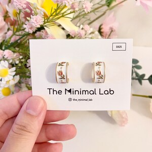 May include: A pair of white hoop earrings featuring a delicate floral pattern with small red tulips and gold accents. The earrings are displayed on a white card labeled S925 and The Minimal Lab, with a small cat icon integrated into the brand name.