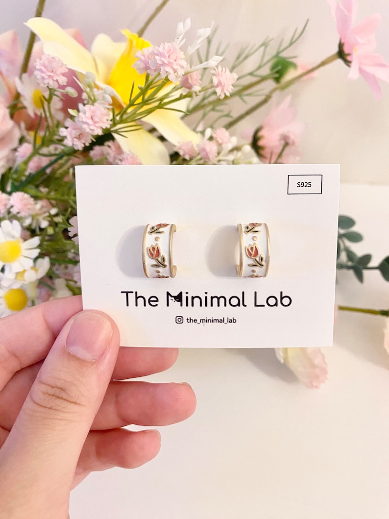 May include: A pair of white hoop earrings featuring a delicate floral pattern with small red tulips and gold accents. The earrings are displayed on a white card labeled S925 and The Minimal Lab, with a small cat icon integrated into the brand name.