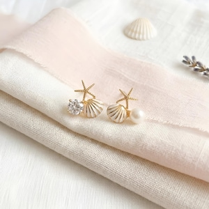 May include: A pair of gold-tone stud earrings featuring a small starfish, a white shell, and a round accent. One earring includes a clear faceted stone, while the other features a smooth white pearl. The earrings are crafted with metallic gold-tone hardware.