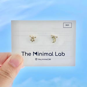 May include: A pair of gold-tone stud earrings featuring a star and a crescent moon, each accented with a small clear baguette stone. The earrings are displayed on a white card labeled The Minimal Lab with an S925 stamp in the corner.