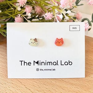 Op de afbeelding: Een paar oorknopjes in de vorm van kattengezichten op een witte displaykaart met het label S925 en The Minimal Lab. De ene oorbel is een crème en bruine lapjeskat, de andere een oranje gestreepte kat. De kaart bevat een Instagram-gebruikersnaam.