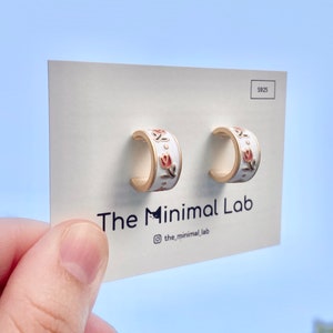 May include: A pair of gold-toned hoop earrings with a white enamel finish and a pink tulip floral pattern. The earrings are mounted on a white display card featuring the text The Minimal Lab and the S925 stamp in the upper right corner.