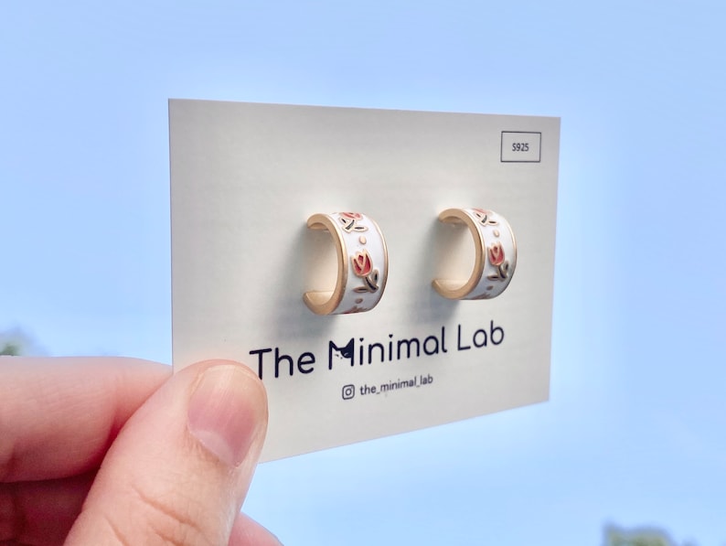 May include: A pair of gold-toned hoop earrings with a white enamel finish and a pink tulip floral pattern. The earrings are mounted on a white display card featuring the text The Minimal Lab and the S925 stamp in the upper right corner.