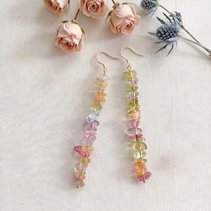 Pode incluir: Um par de brincos longos pendentes com fios de pedras preciosas multicoloridas em tons de rosa, verde, amarelo e roxo. Os brincos estão suspensos em ganchos de metal dourado e são exibidos ao lado de rosas cor-de-rosa secas e cardos azuis.