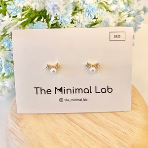 Puede incluir: Un par de pendientes de oro con una peque&ntilde;a perla y un dise&ntilde;o de lazo. Los pendientes se muestran en una tarjeta blanca con el texto "The Minimal Lab" y "@the_minimal_lab".