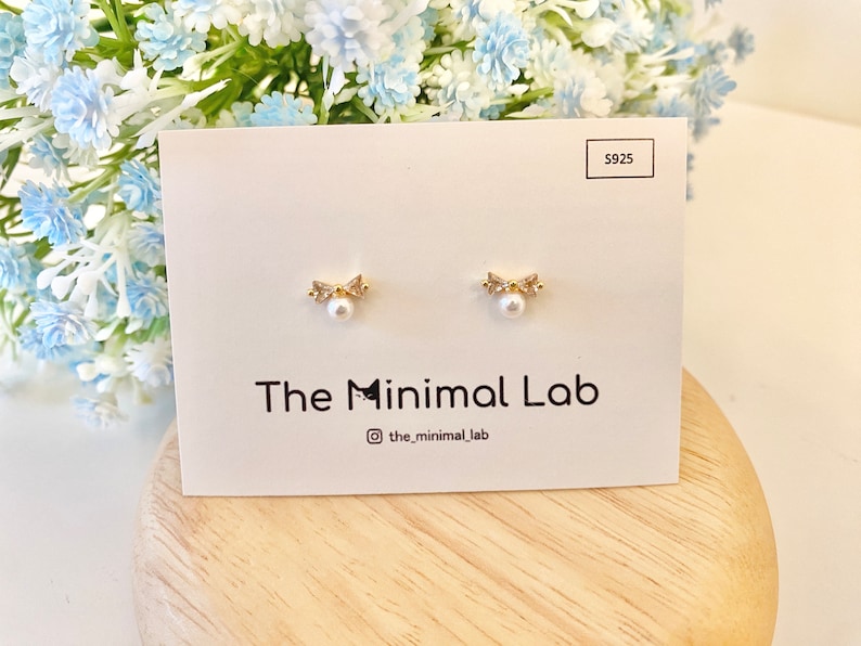 Puede incluir: Un par de pendientes de oro con una peque&ntilde;a perla y un dise&ntilde;o de lazo. Los pendientes se muestran en una tarjeta blanca con el texto "The Minimal Lab" y "@the_minimal_lab".
