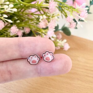 May include: A pair of small stud earrings featuring a cat paw print design. Each earring has a translucent base with a light pink central pad and four smaller pink toe pads, all outlined in a thin brown border with a smooth, glossy finish.