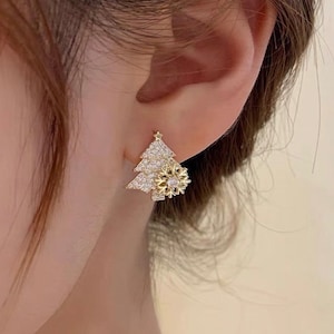 May include: A gold-toned Christmas tree stud earring with a sparkling snowflake accent. The tree is encrusted with small, clear crystals, and the snowflake has a detailed, openwork design. The earring is shown on an ear.