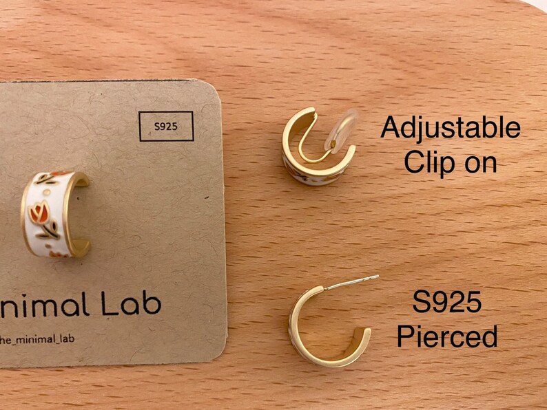 May include: Gold-toned hoop earrings featuring a white enamel surface with small orange and green floral motifs. The set includes an adjustable clip-on earring and a pierced earring with an S925 post, displayed on a tan card labeled Minimal Lab.