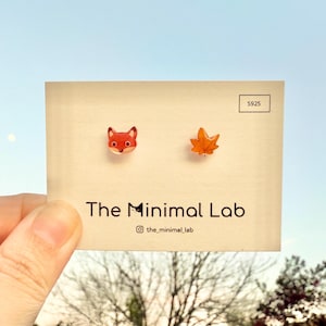 Puede incluir: Un par de pendientes con un pequeño zorro y una hoja de arce. Los pendientes se muestran en una tarjeta con el texto "The Minimal Lab" y el nombre de usuario de Instagram "@the_minimal_lab".