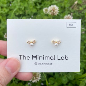 Puede incluir: Un par de pendientes de oro con una perla y un peque&ntilde;o dise&ntilde;o de lazo. Los pendientes est&aacute;n en una tarjeta blanca con el texto "The Minimal Lab" y "@the_minimal_lab".