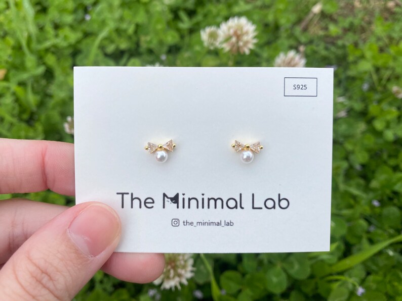 Puede incluir: Un par de pendientes de oro con una perla y un peque&ntilde;o dise&ntilde;o de lazo. Los pendientes est&aacute;n en una tarjeta blanca con el texto "The Minimal Lab" y "@the_minimal_lab".
